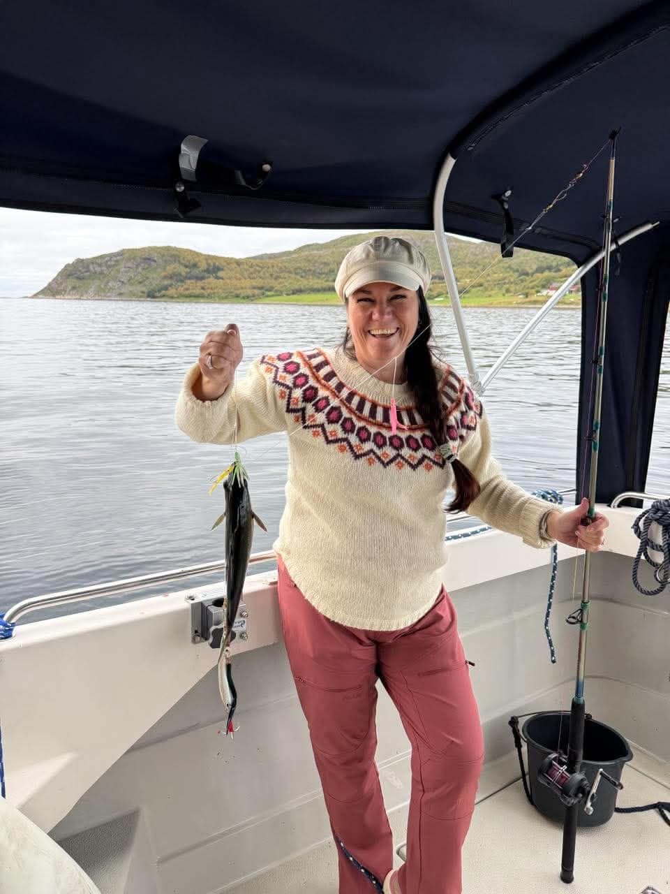 A smiling guest in a knitted jumper holding up her catch on the line, the boat's canopy above and the island behind