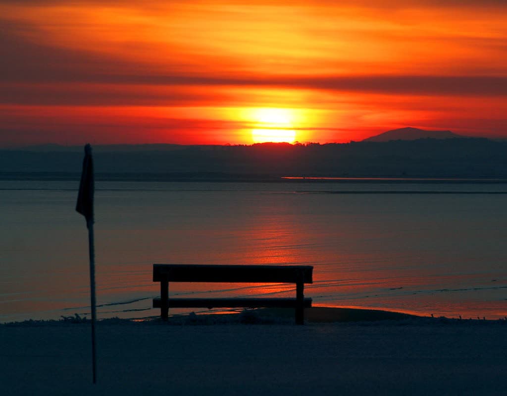 A flag on a green by the sea, a bench beside it and a low orange sun over the water