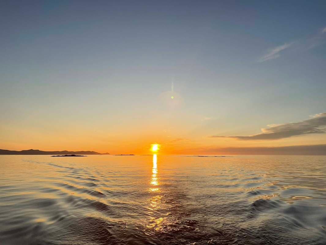 The midnight sun low over a calm sea, the boat's wake catching the light