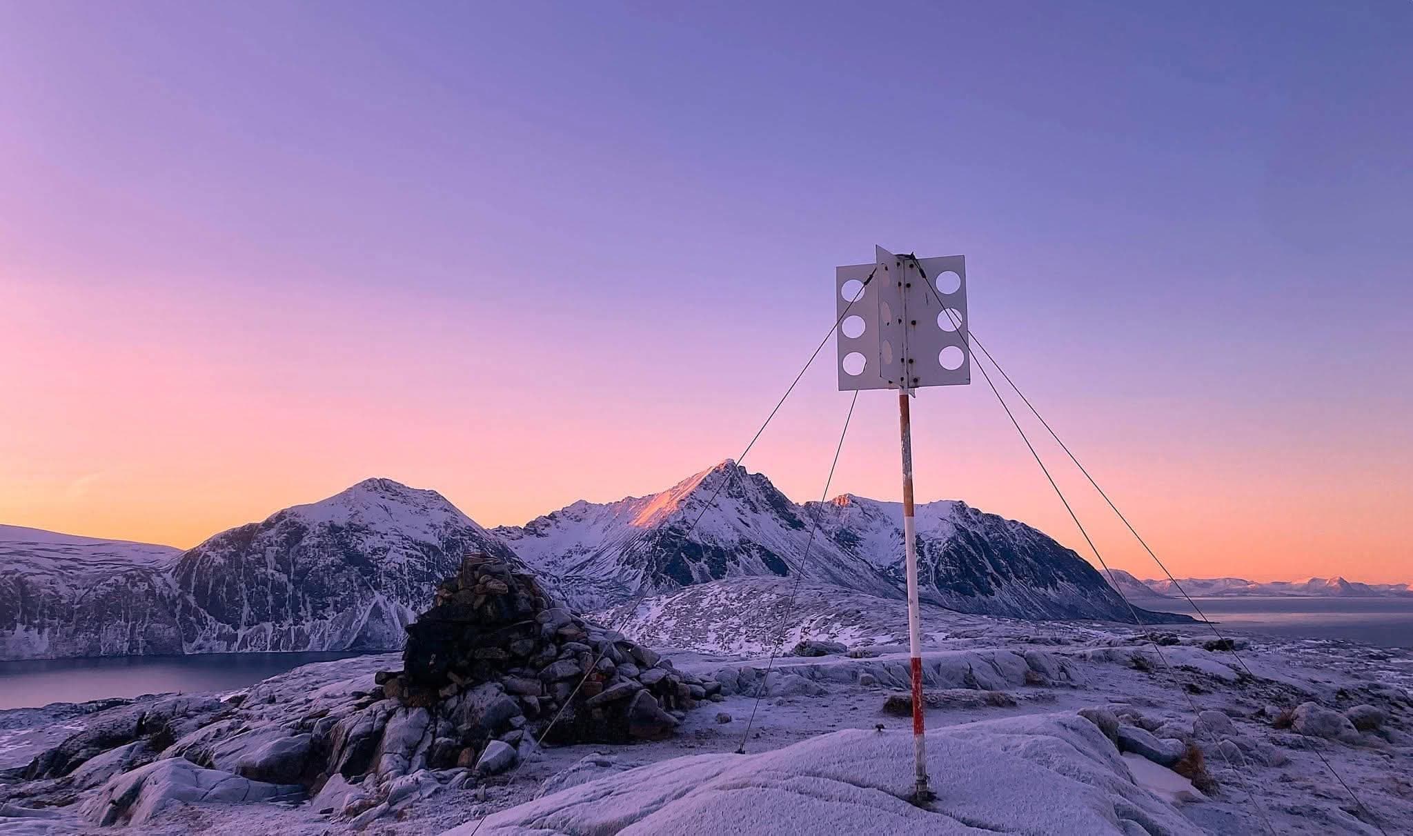 A cairn and a survey mast on a frosted summit, snow-covered peaks glowing pink under a violet winter sky